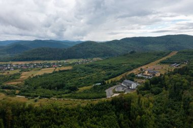 Yoğun çam ormanlarıyla kaplı katmanlı dağ sıralarının ve bulutlu gökyüzünün altındaki vadideki küçük kasabanın çarpıcı manzarası.