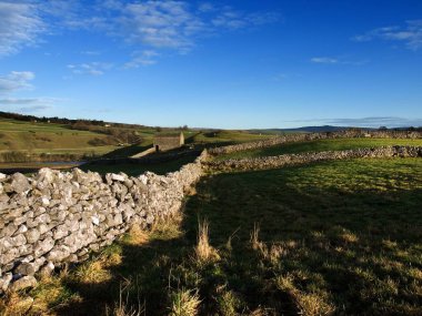 Kırsal evlerin, eski taş duvarların ve güneşli mavi gökyüzünün altındaki tarım arazilerinin resimsel manzarası.