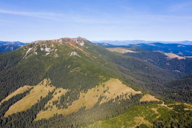Güneşli bir günde mavi gökyüzünün altında yoğun çam ormanı ve yeşil vadi ile kaplı katmanlı sıradağların çarpıcı manzarası.