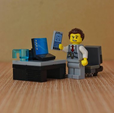 Oyuncak bir erkek figürünün yakın plan fotoğrafı, evden çalışıyor ve bir telefon tutuyor, masada bilgisayarın yanında duruyor, takım elbise ve kravat takıyor.