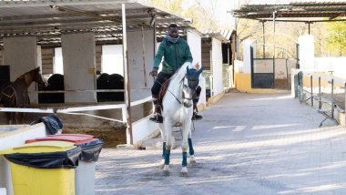 Çiftlikte beyaz bir ata binen, kalın giysili, yetişkin siyah bir erkek.