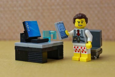 Oyuncak bir erkek figürünün yakın plan fotoğrafı, evden çalışıyor ve bir telefon tutuyor, masanın üstünde bir bilgisayarın yanında duruyor, şort ve kravat takıyor.