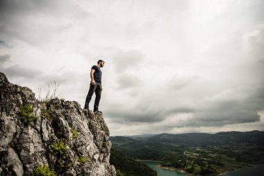 Uçurumun kenarında duran bir adam güzel manzaraları ve arka planda bulutlu gökyüzü olan.