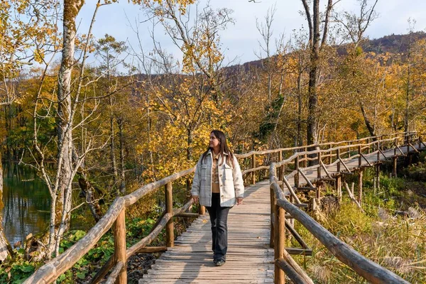 一位年轻女子走在一座木桥上 欣赏着美丽的黄秋森林 — 图库照片