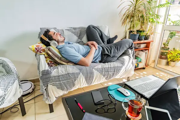 stock image A young male in sweatpants laying on a sofa at home listening to music with headphones