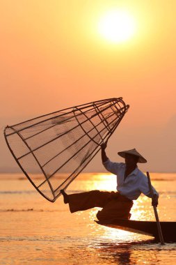 Gölün kenarında bir balıkçının silueti günbatımında el yapımı bir ağla geleneksel bir şekilde balık yakalıyor.