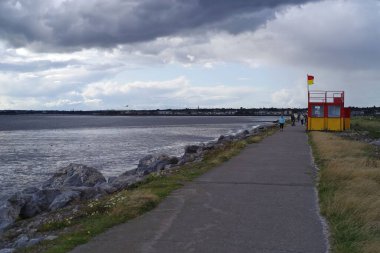 İrlanda, Dublin 'deki Bull Adası' nda bulunan Dollymount Strand 'da insanlar deniz kıyısında yürüyor. Cankurtaran yolda dursun. Dramatik bulutlar.