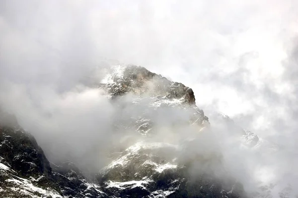 雪と雲に覆われた岩だらけの山の頂上は暗く寒い天候で — ストック写真