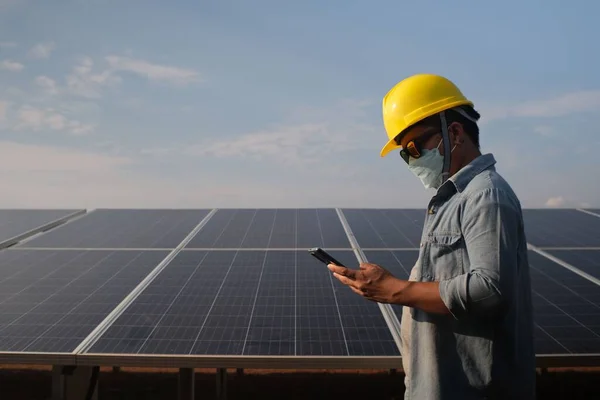 一名戴着黄色头盔和面罩的承包商在太阳能电池板的背景下按下了手机 — 图库照片