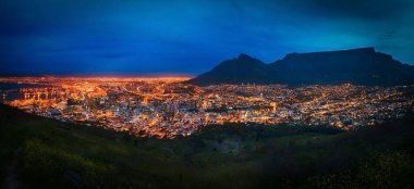 Aydınlatılmış şehir manzarasının panoramik bir görüntüsü. Arka planda mavi bulutlu gökyüzünün altında dağ var.
