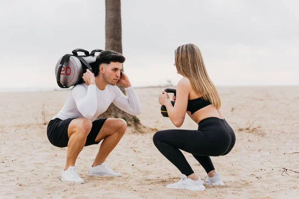 stock image A young athletic male with a female doing back squats using a sandbag and a kettlebell