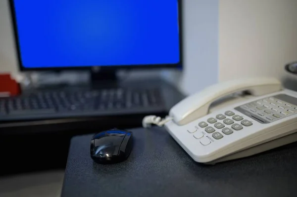 桌上有一个黑色鼠标和一个白色电话 背景上有一个蓝色的监视器屏幕和一个键盘 浅水区深度 — 图库照片
