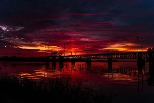 橋の上に抽象的な雲と美しい緋色の夕日の空の風景 水面に反射する波の海 — ストック写真