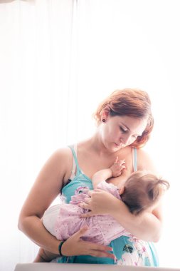 Güneşli bir odada bebeğini emziren genç bir anne.