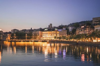 Zürih 'in aydınlattığı akşam binalar mavi gökyüzünün altındaki durgun suyu yansıtıyordu.