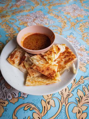 Bir porsiyon sıcak roti canai ile bir kase köri ve desenli masa örtüsü.