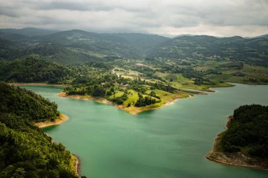 Sırbistan 'ın batısında gri bulutlu gökyüzü altında yeşil bitki örtüsüyle çevrili Rovni Gölü' nün hava manzarası