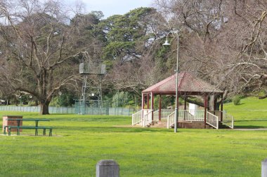 Güzel ahşap bir çadırı ve yeşil bir bankı olan yeşil bir park manzarası.