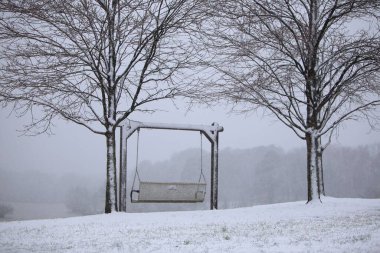 Parkta sisli bir kış günü ve iki kuru ağaç arasında karla kaplı bir salıncak.