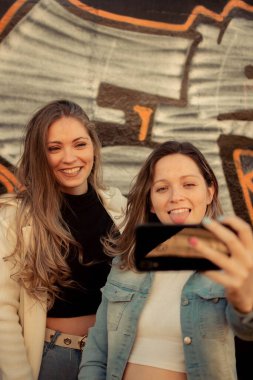 İki bayan arkadaşın gülümseyerek ve sokakta selfie çekerken çekilmiş dikey bir fotoğrafı.