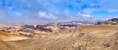 Çölün panoramik bir görüntüsü, dalgalı kum tepeleri ve mavi gökyüzünün altındaki kayalık tepelerin olduğu çorak bir bölge.