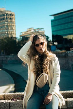 Açık renk ceketli, güneş gözlüklü genç bir kadının dikey görüntüsü. Sokakta alçak bir taş duvarda oturuyor.