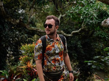 Çiçekli gömlekli, siyah güneş gözlüklü, arka planda yeşil ağaçlarla poz veren beyaz bir adam.