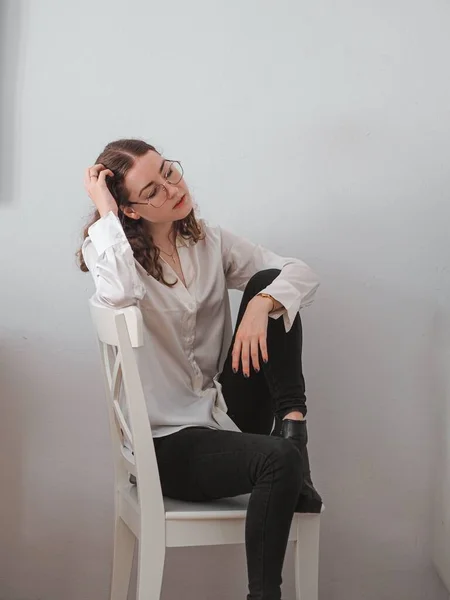 stock image A vertical shot of a woman posing on the wooden chair on the background of a white wall