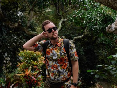 Çiçekli gömlekli, siyah güneş gözlüklü, arka planda yeşil ağaçlarla poz veren beyaz bir adam.