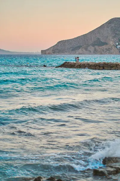 在被迷人的蓝色波浪形大海环绕着的岩石路上站着的一个垂直的人 — 图库照片