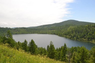 Bulutlu bir günde tepe ve ormanlarla çevrili bir göl manzarası.