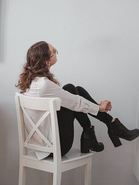 A vertical shot of a woman posing on the wooden chair on the background of a white wall