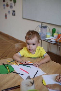 Masada oturan ve anaokulunda resim çizen beyaz bir çocuk.