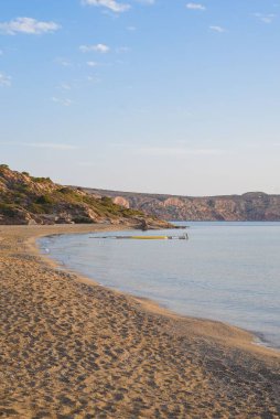 Mavi gökyüzünün altında sakin bir deniz ve uçurumun olduğu boş bir sahilin dikey görüntüsü.
