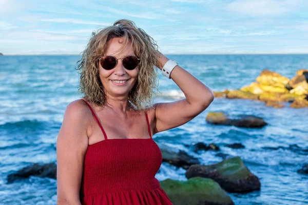 50代の女性が赤いドレスと黒のサングラスをかけて海に向かって微笑んだ — ストック写真