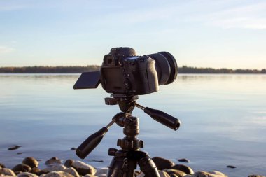 Sakin bir gölün kayalık sahilinde, tripodun üzerinde profesyonel bir kameraya yakın çekim.
