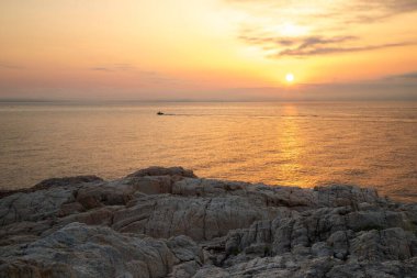 Kayalık bir uçurumun üzerinde yakın çekim ve sakin denizin yüzeyini boyayan batan güneş.
