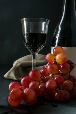 Masada bir kadeh kırmızı şarap, şarap şişesi ve bir demet üzüm var.