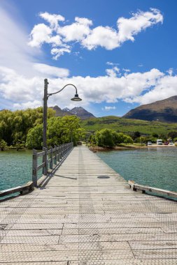 Glenorchy 'deki ahşap bir iskelenin dikey görüntüsü. Arka planda bulutlu mavi bir gökyüzü var. Otago, Yeni Zelanda.