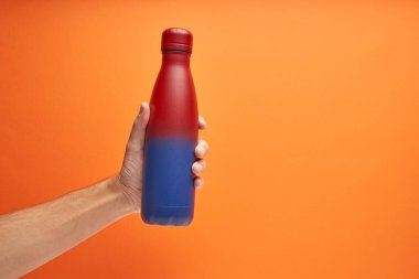 Turuncu arka planda kırmızı ve mavi bir şişe tutan beyaz bir el.