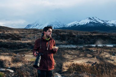 Profesyonel kameralı genç bir adam karlı dağların arkasında sandviç yiyor.