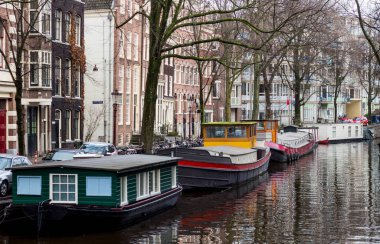 Amsterdam 'da nehir kenarına yanaşan teknelerin güzel bir görüntüsü. Arka planda ağaçlar ve mimariler var.