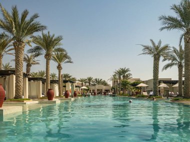 Al Wathba Desert Resort ve Spa 'da palmiye ağaçları ve kabinlerle dolu uçsuz bucaksız sonsuz bir havuz.