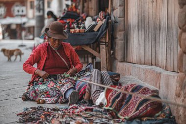 Peru, Cusco 'da geleneksel kıyafetleriyle yerde oturan ve el yapımı kumaşlar yapan bir kadın.