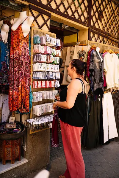 女性の観光客の笑顔の垂直ショットと服市場でラック上のアクセサリーを選択 — ストック写真