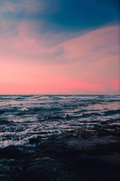 A vertical shot of a seascape under pink and blue clouds in the sky
