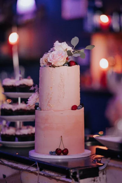 一个垂直拍摄的婚礼蛋糕 装饰着粉红色的玫瑰和水果 背景上有彩灯 — 图库照片
