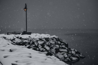 Karla kaplı kayalık bir sahilde sisli bir kış gününde bankı aydınlatan bir lambanın manzarası.