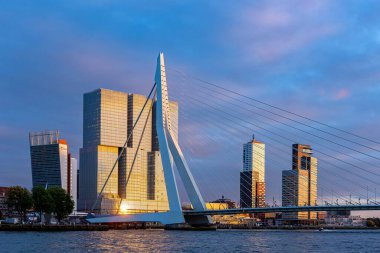 Warm sun reflection on Erasmus bridge and high rise buildings of the financial district in the Dutch city in the background against a sunset sky clipart