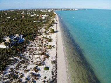 Mavi sularla çevrili bir sahil, ağaçlarla çevrili kumlu evler ve mavi bir gökyüzü.